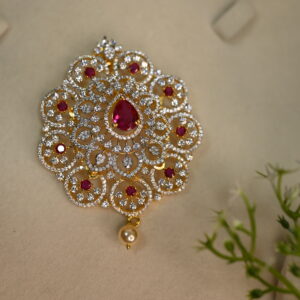 The image shows a beautiful pendant. It appears to be made of gold and is adorned with numerous small, colorless stones, likely cubic zirconia, and a larger, central red stone. The pendant has a delicate, flower-like design with intricate patterns. It also has a small, pearl-like drop hanging from the bottom.