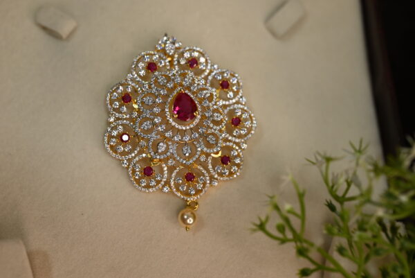 The image shows a beautiful pendant. It appears to be made of gold and is adorned with numerous small, colorless stones, likely cubic zirconia, and a larger, central red stone. The pendant has a delicate, flower-like design with intricate patterns. It also has a small, pearl-like drop hanging from the bottom.