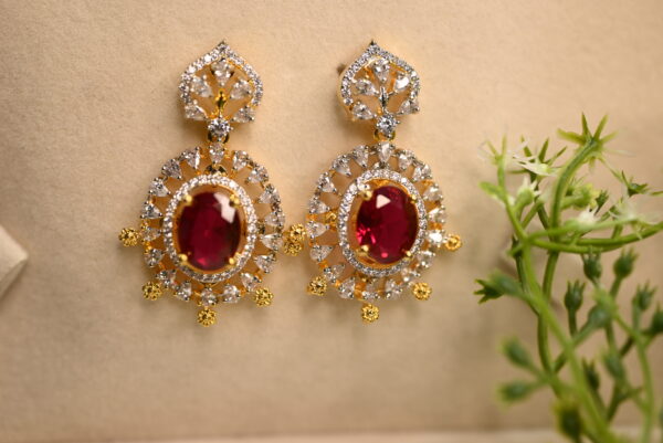 Image: A pair of elegant earrings are showcased on a beige surface, adorned with a delicate sprig of greenery in the corner. Earrings: Style: Drop earrings with a classic, ornate design. Color: The earrings are primarily gold-toned. CZ Earring