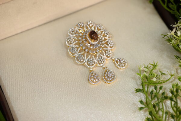 The image shows a beautiful, ornate piece of jewelry. It appears to be a brooch or pendant, with a large, round, brown gemstone in the center. The gemstone is surrounded by a delicate, intricate design of smaller stones, which might be diamonds or other precious stones. The design is in a gold color, possibly gold or gold-plated. The jewelry is displayed on a luxurious, cream-colored surface, suggesting that it is a high-quality, valuable piece. There are also some green and white flowers in the background, adding a touch of natural beauty to the image.
