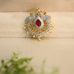 The image shows a beautiful piece of jewelry, likely a pendant or brooch. It has a central teardrop-shaped red stone, surrounded by a cluster of smaller white stones. The design is intricate, with delicate goldwork forming a floral pattern around the stones. The jewelry is placed on a soft, beige surface, possibly velvet or satin, and a sprig of green leaves adds a touch of nature to the scene. The overall impression is one of elegance and luxury.