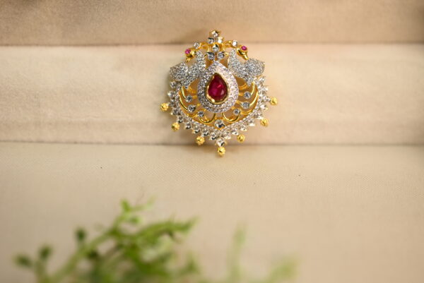 The image shows a beautiful piece of jewelry, likely a pendant or brooch. It has a central teardrop-shaped red stone, surrounded by a cluster of smaller white stones. The design is intricate, with delicate goldwork forming a floral pattern around the stones. The jewelry is placed on a soft, beige surface, possibly velvet or satin, and a sprig of green leaves adds a touch of nature to the scene. The overall impression is one of elegance and luxury.