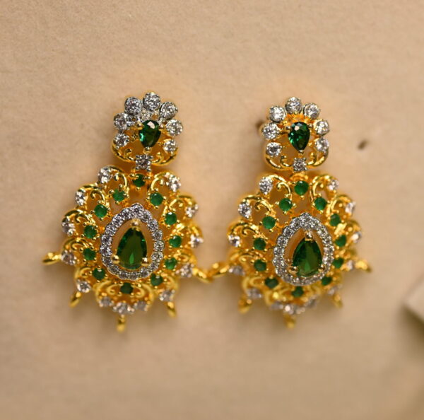 The image shows a pair of elegant earrings. They are made of gold and adorned with sparkling white and green CZ diamonds. The design is intricate, featuring a floral motif with delicate filigree work. The earrings have a drop style, with the diamonds cascading downwards. The overall effect is one of luxury and sophistication.