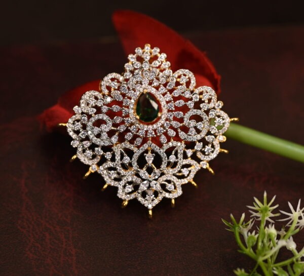 The image shows a gold-toned pendant resting on a beige surface. The pendant features a large, oval-shaped green gemstone at its center. Surrounding the gemstone is a delicate, intricate design of smaller, white stones, likely cubic zirconia (CZ), creating a floral or lacy pattern. The pendant has a small, pointed base.