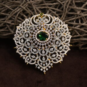 The image shows a gold-toned pendant resting on a beige surface. The pendant features a large, oval-shaped green gemstone at its center. Surrounding the gemstone is a delicate, intricate design of smaller, white stones, likely cubic zirconia (CZ), creating a floral or lacy pattern. The pendant has a small, pointed base.
