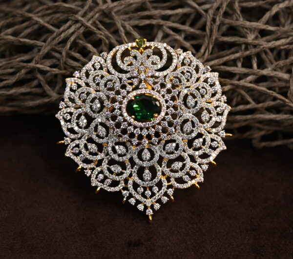 The image shows a gold-toned pendant resting on a beige surface. The pendant features a large, oval-shaped green gemstone at its center. Surrounding the gemstone is a delicate, intricate design of smaller, white stones, likely cubic zirconia (CZ), creating a floral or lacy pattern. The pendant has a small, pointed base.