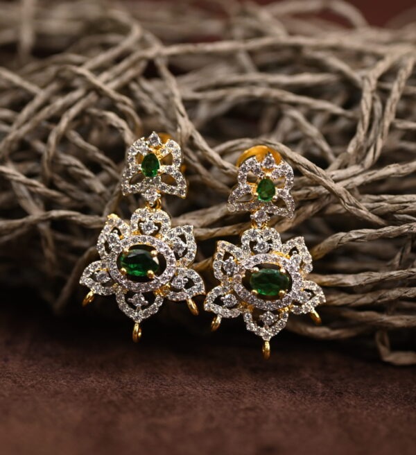 The background of the image is a neutral color, likely to highlight the earrings. There appears to be a small sprig of greenery in the bottom right corner of the image, adding a natural touch.