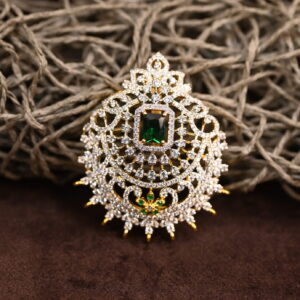 The image shows a gold-toned pendant resting on a beige surface. The pendant features a large, oval-shaped green gemstone at its center. Surrounding the gemstone is a delicate, intricate design of smaller, white stones, likely cubic zirconia (CZ), creating a floral or lacy pattern. The pendant has a small, pointed base.