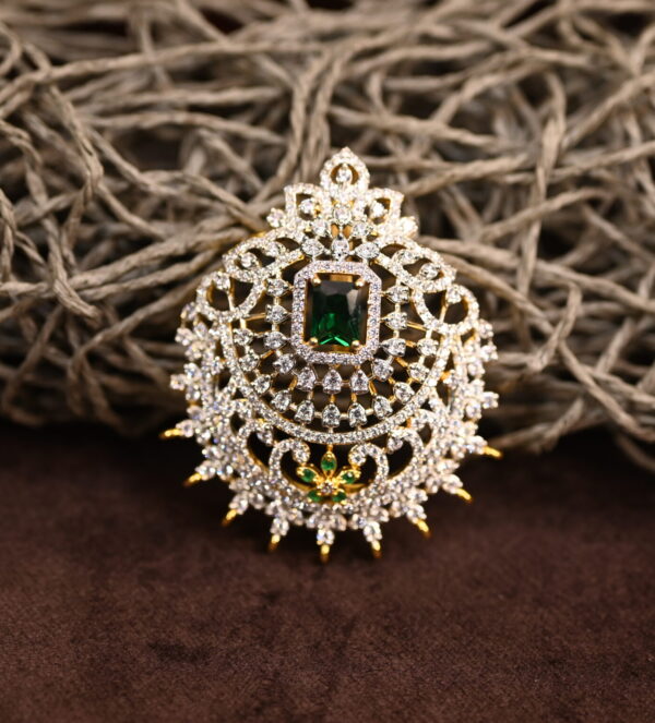 The image shows a gold-toned pendant resting on a beige surface. The pendant features a large, oval-shaped green gemstone at its center. Surrounding the gemstone is a delicate, intricate design of smaller, white stones, likely cubic zirconia (CZ), creating a floral or lacy pattern. The pendant has a small, pointed base.
