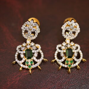 The background of the image is a neutral color, likely to highlight the earrings. There appears to be a small sprig of greenery in the bottom right corner of the image, adding a natural touch.