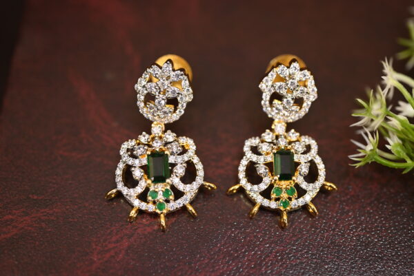 The background of the image is a neutral color, likely to highlight the earrings. There appears to be a small sprig of greenery in the bottom right corner of the image, adding a natural touch.