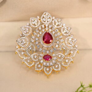 The image shows a close-up of a gold-toned pendant. The pendant has a large, teardrop-shaped Pink stone in the center, likely a cubic zirconia simulating a ruby. This central stone is surrounded by a halo of smaller white cubic zirconia stones. The pendant is further adorned with intricate filigree work, creating a delicate and ornate frame.
