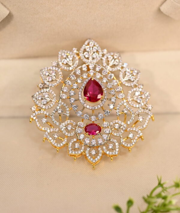 The image shows a close-up of a gold-toned pendant. The pendant has a large, teardrop-shaped Pink stone in the center, likely a cubic zirconia simulating a ruby. This central stone is surrounded by a halo of smaller white cubic zirconia stones. The pendant is further adorned with intricate filigree work, creating a delicate and ornate frame.