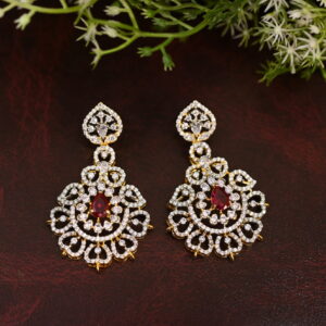 The image shows a close-up of a pair of earrings. The earrings have a circular or floral shape with a dangling drop element. They are made of gold-toned metal and adorned with sparkling white stones. There is a prominent red stone in the center of each earring. The earrings are resting on a dark brown surface with a leafy green plant in the background.
