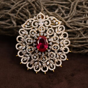 The image shows a beautiful pendant adorned with a large red gemstone, likely a ruby, surrounded by a halo of sparkling white diamonds. The pendant is set in a gold-toned metal and has a delicate, intricate design. It appears to be resting on a bed of brown, textured material.
