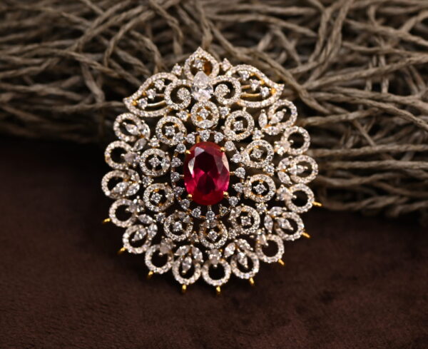 The image shows a beautiful pendant adorned with a large red gemstone, likely a ruby, surrounded by a halo of sparkling white diamonds. The pendant is set in a gold-toned metal and has a delicate, intricate design. It appears to be resting on a bed of brown, textured material.