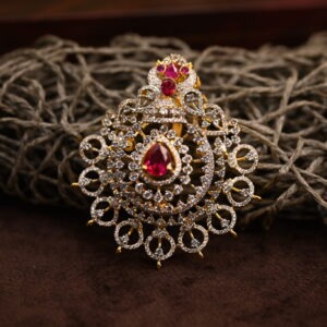 The image shows a close-up of a gold-toned pendant. The pendant has a large, teardrop-shaped pink stone in the center, likely a cubic zirconia simulating a ruby. This central stone is surrounded by a halo of smaller white cubic zirconia stones. The pendant is further adorned with intricate filigree work, creating a delicate and ornate frame.
