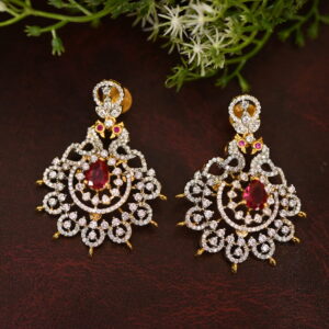 The image shows a pair of elegant earrings on a dark brown surface. The earrings are made of gold and feature a circular design with numerous small diamonds. In the center of each earring, there is a prominent red stone, likely a ruby or a similar gemstone. The earrings are adorned with delicate floral patterns, adding to their intricate design. The background is a dark brown surface, possibly leather, with some green foliage adding a touch of nature.