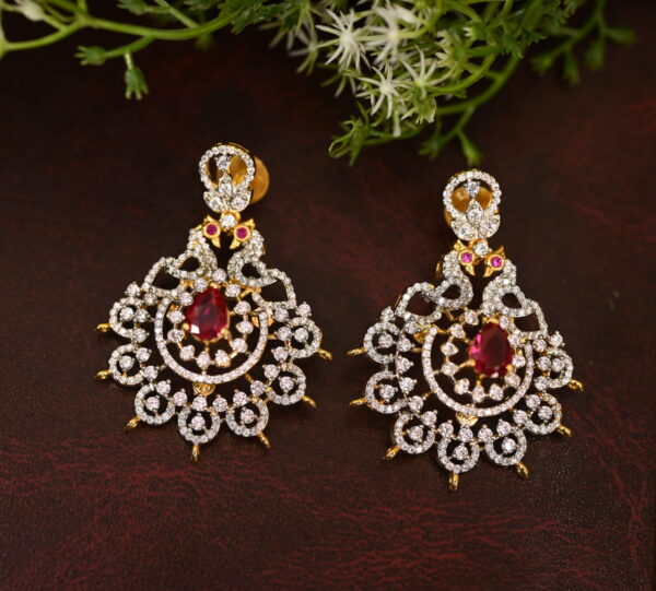 The image shows a pair of elegant earrings on a dark brown surface. The earrings are made of gold and feature a circular design with numerous small diamonds. In the center of each earring, there is a prominent red stone, likely a ruby or a similar gemstone. The earrings are adorned with delicate floral patterns, adding to their intricate design. The background is a dark brown surface, possibly leather, with some green foliage adding a touch of nature.