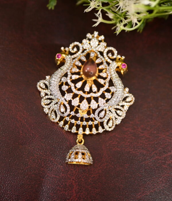 The image shows a beautiful CZ pendant featuring a pair of peacocks with a large, round ruby in the center. The peacocks are adorned with white CZ stones, creating a dazzling and intricate design. A small, bell-shaped pendant hangs from the bottom, adding a touch of elegance. The overall effect is a luxurious and eye-catching piece of jewelry.