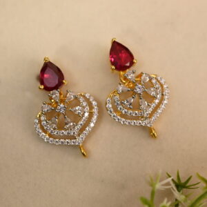 The image shows a pair of elegant earrings. Each earring features a heart-shaped design crafted from gold-toned metal. The hearts are intricately embellished with a cluster of sparkling white cubic zirconia (CZ) stones. Dangling from the bottom of each heart is a single, teardrop-shaped red CZ stone. The earrings are displayed on a light-colored surface with a small sprig of greenery in the background.