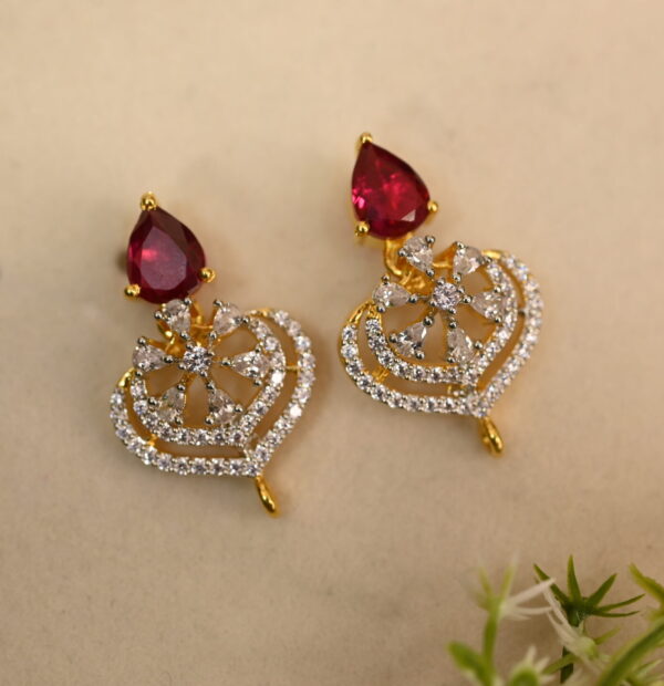 The image shows a pair of elegant earrings. Each earring features a heart-shaped design crafted from gold-toned metal. The hearts are intricately embellished with a cluster of sparkling white cubic zirconia (CZ) stones. Dangling from the bottom of each heart is a single, teardrop-shaped red CZ stone. The earrings are displayed on a light-colored surface with a small sprig of greenery in the background.