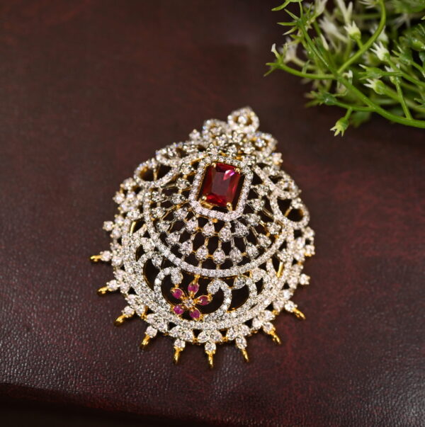 The image shows a close-up of a delicate pendant resting on a dark, textured surface. The pendant has a circular design with intricate detailing and a large, square-cut, red gemstone set in the center. The gemstone is surrounded by a halo of smaller, white stones, likely cubic zirconia.