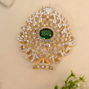 The image shows a gold-toned pendant resting on a beige surface. The pendant features a large, oval-shaped green gemstone at its center. Surrounding the gemstone is a delicate, intricate design of smaller, white stones, likely cubic zirconia (CZ), creating a floral or lacy pattern. The pendant has a small, pointed base.