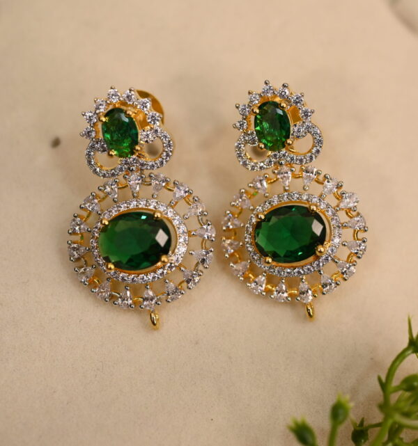 The background of the image is a neutral color, likely to highlight the earrings. There appears to be a small sprig of greenery in the bottom right corner of the image, adding a natural touch.