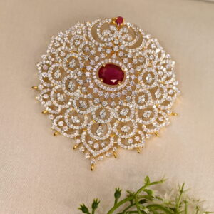 The image shows a close-up of a gold-toned pendant. The pendant has a large, teardrop-shaped red stone in the center, likely a cubic zirconia simulating a ruby. This central stone is surrounded by a halo of smaller white cubic zirconia stones. The pendant is further adorned with intricate filigree work, creating a delicate and ornate frame.