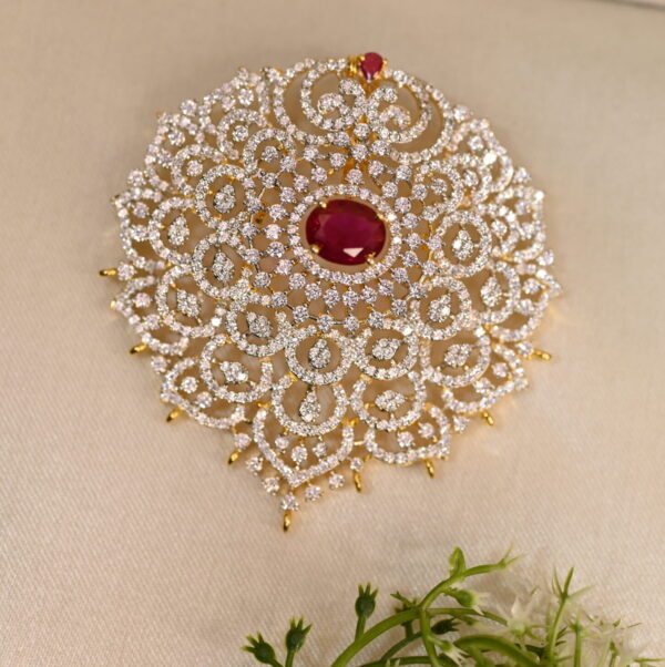 The image shows a close-up of a gold-toned pendant. The pendant has a large, teardrop-shaped red stone in the center, likely a cubic zirconia simulating a ruby. This central stone is surrounded by a halo of smaller white cubic zirconia stones. The pendant is further adorned with intricate filigree work, creating a delicate and ornate frame.