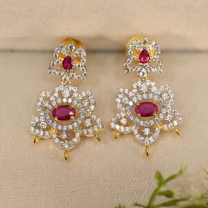 The image shows a pair of elegant earrings. They are made of gold and feature a cluster of cubic zirconia (CZ) stones in the center. The CZ stones are surrounded by smaller diamonds, creating a dazzling effect. The earrings have a delicate and intricate design with a dangling drop shape. They are resting on a brown surface, possibly leather or velvet, with a small sprig of greenery in the background.