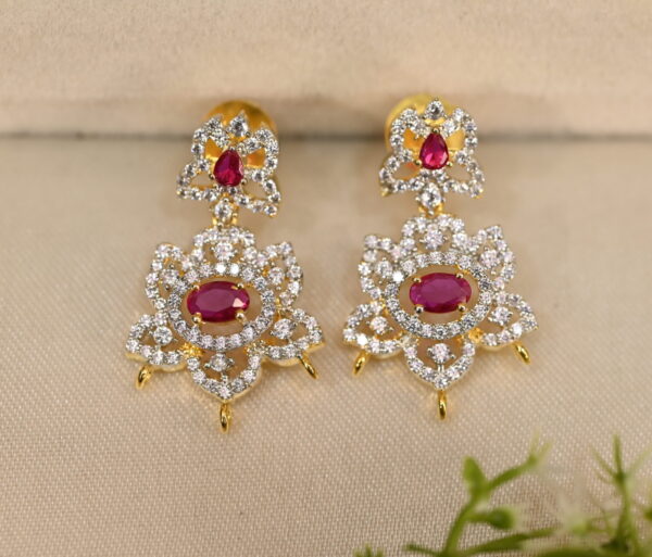 The image shows a pair of elegant earrings. They are made of gold and feature a cluster of cubic zirconia (CZ) stones in the center. The CZ stones are surrounded by smaller diamonds, creating a dazzling effect. The earrings have a delicate and intricate design with a dangling drop shape. They are resting on a brown surface, possibly leather or velvet, with a small sprig of greenery in the background.