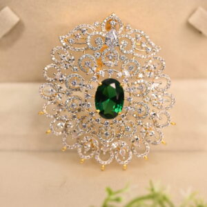 The image shows a gold-toned pendant resting on a beige surface. The pendant features a large, oval-shaped green gemstone at its center. Surrounding the gemstone is a delicate, intricate design of smaller, white stones, likely cubic zirconia (CZ), creating a floral or lacy pattern. The pendant has a small, pointed base.