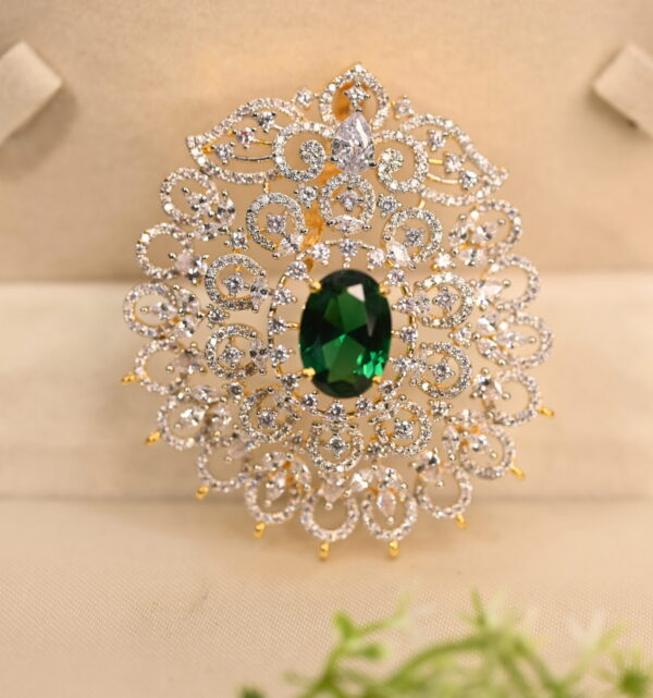 The image shows a gold-toned pendant resting on a beige surface. The pendant features a large, oval-shaped green gemstone at its center. Surrounding the gemstone is a delicate, intricate design of smaller, white stones, likely cubic zirconia (CZ), creating a floral or lacy pattern. The pendant has a small, pointed base.