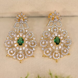 The background of the image is a neutral color, likely to highlight the earrings. There appears to be a small sprig of greenery in the bottom right corner of the image, adding a natural touch.