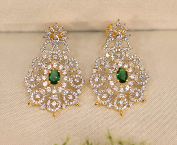 The background of the image is a neutral color, likely to highlight the earrings. There appears to be a small sprig of greenery in the bottom right corner of the image, adding a natural touch.