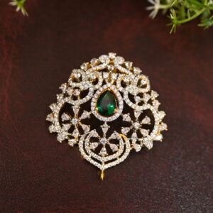 The image shows a gold-toned pendant resting on a beige surface. The pendant features a large, oval-shaped green gemstone at its center. Surrounding the gemstone is a delicate, intricate design of smaller, white stones, likely cubic zirconia (CZ), creating a floral or lacy pattern. The pendant has a small, pointed base.
