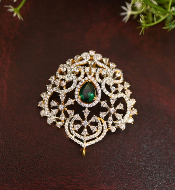 The image shows a gold-toned pendant resting on a beige surface. The pendant features a large, oval-shaped green gemstone at its center. Surrounding the gemstone is a delicate, intricate design of smaller, white stones, likely cubic zirconia (CZ), creating a floral or lacy pattern. The pendant has a small, pointed base.