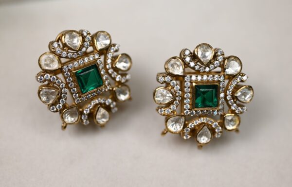 The image shows a pair of earrings. They have a square-shaped center with a large green gemstone in the middle. Surrounding the green gemstone are smaller white gemstones arranged in a circular pattern. The earrings are made of gold.