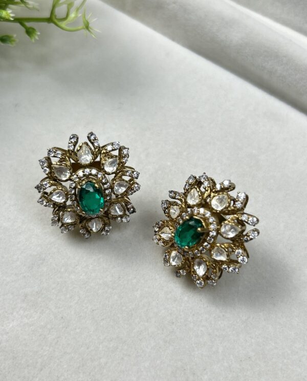 "Close-up of a pair of elegant gold stud earrings with a delicate floral design. Each earring is adorned with a cluster of sparkling moissanites surrounding a captivating emerald-cut green gemstone."