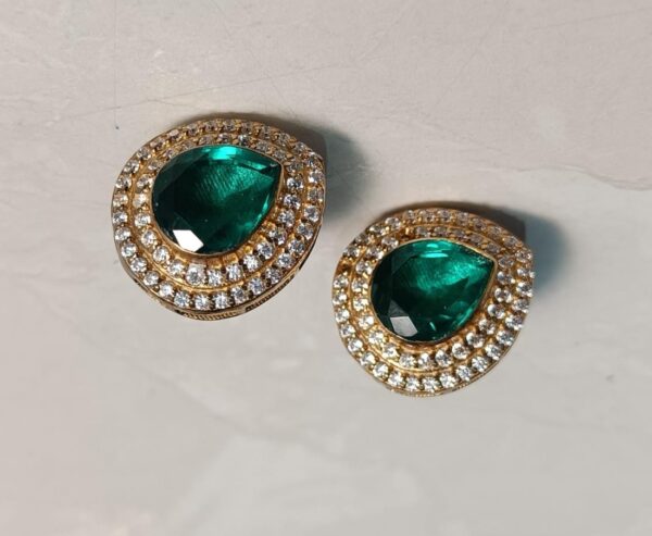 The image shows a pair of elegant earrings. Each earring features a large, pear-shaped emerald-green gemstone as the centerpiece. The gemstones are surrounded by a halo of smaller, round-cut white stones, creating a dazzling frame. The earrings appear to be set in a warm-toned metal, possibly gold or brass.
