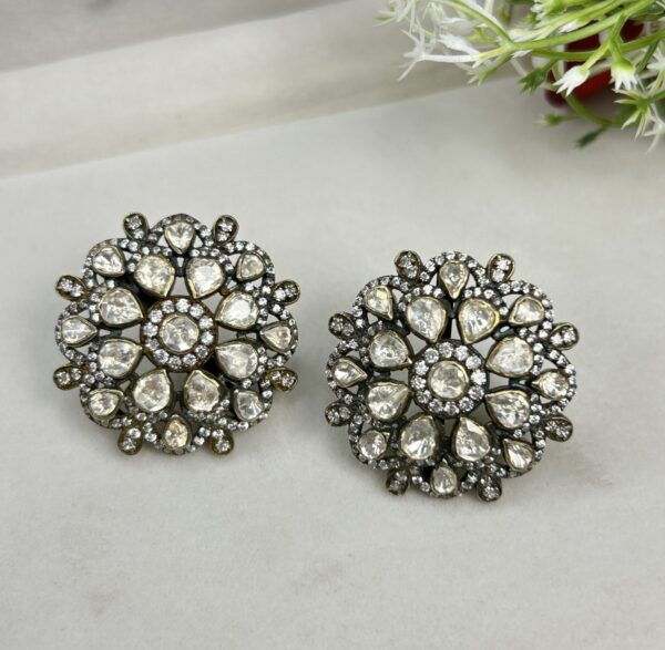 The image shows a pair of round-shaped stud earrings with a floral design. The earrings are made of white moissanite stones, set in a silver-toned metal. The floral design has a central motif with smaller stones surrounding it. The earrings have a vintage or antique look to them.