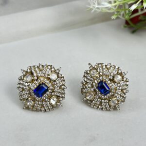 The image shows a pair of elegant earrings. Each earring features a square-shaped design with a central, square-cut blue gemstone, likely a sapphire or a blue moissanite. Surrounding the central stone is a delicate floral pattern made up of smaller, white gemstones, likely diamonds or white moissanites. The background of the image is a light, neutral color, possibly a marble or a light wood surface.