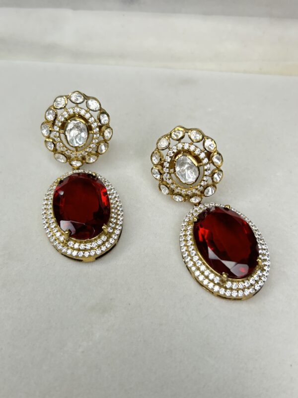The image shows a pair of elegant dangle earrings. Each earring features a large, oval-shaped red gemstone surrounded by a halo of smaller white gemstones. The top of each earring has a circular design with multiple layers of white gemstones.