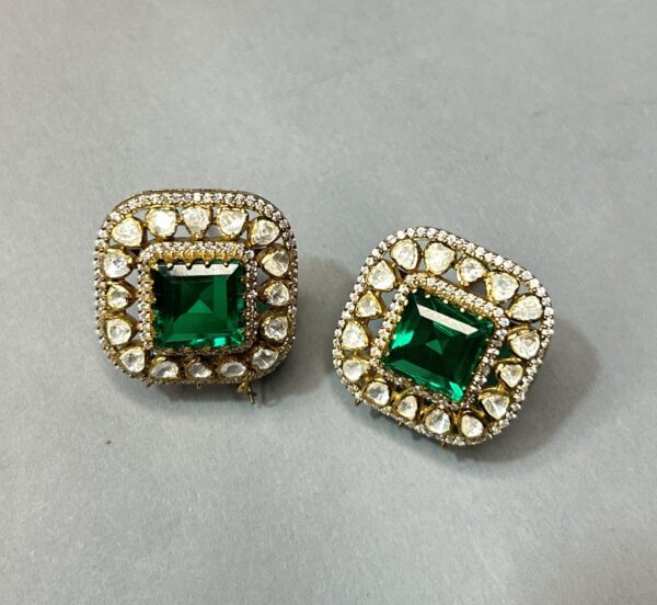 The image shows a pair of stunning earrings. They are made of gold and feature a large, square-cut emerald gemstone in the center. The emerald is surrounded by a halo of smaller, white gemstones, possibly diamonds. The earrings have a delicate, intricate design with a floral pattern. The overall effect is one of elegance and luxury.