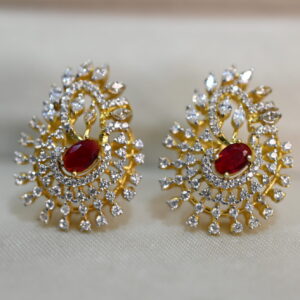 The image shows a pair of earrings. They have a gold-tone setting and are adorned with numerous small, colorless stones, likely diamonds or cubic zirconia, arranged in a swirling pattern. In the center of each earring, there is a larger, round red stone, possibly a ruby or a synthetic ruby. The earrings seem to be inspired by the design of a peacock's tail feathers, with the swirling stones mimicking the fan-like shape.