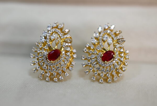 The image shows a pair of earrings. They have a gold-tone setting and are adorned with numerous small, colorless stones, likely diamonds or cubic zirconia, arranged in a swirling pattern. In the center of each earring, there is a larger, round red stone, possibly a ruby or a synthetic ruby. The earrings seem to be inspired by the design of a peacock's tail feathers, with the swirling stones mimicking the fan-like shape.