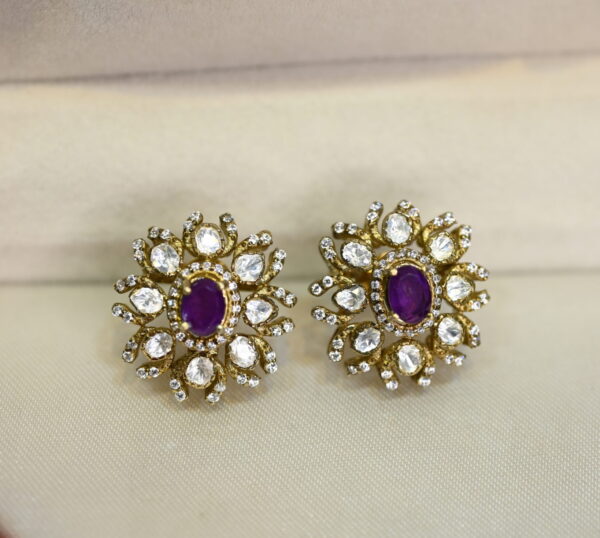 The image shows a pair of stud earrings. The earrings are gold-toned and have a floral design with a purple stone in the center. The design is intricate and detailed, with small white stones surrounding the center stone.