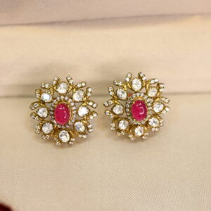 The image shows a pair of stud earrings. The earrings are gold-toned and have a floral design with a purple stone in the center. The design is intricate and detailed, with small white stones surrounding the center stone.